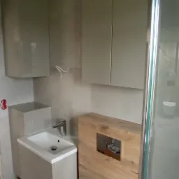 Wnętrze łazienki w trakcie wykańczania, widoczna podwieszana toaleta, umywalka z szafką, zabudowa geberitu z drewnopodobnych paneli, oraz częściowo widoczna kabina prysznicowa.