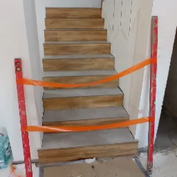 Schody wyłożone płytkami imitującymi drewno, zabezpieczone taśmą ostrzegawczą i poziomnicami, widoczne ślady po pracach wykończeniowych na ścianie.