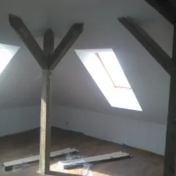 Poddasze z drewnianymi belkami konstrukcyjnymi, oknem dachowym i częściowo ułożoną podłogą z paneli; widoczne opakowania po panelach i ulotka informacyjna producenta.