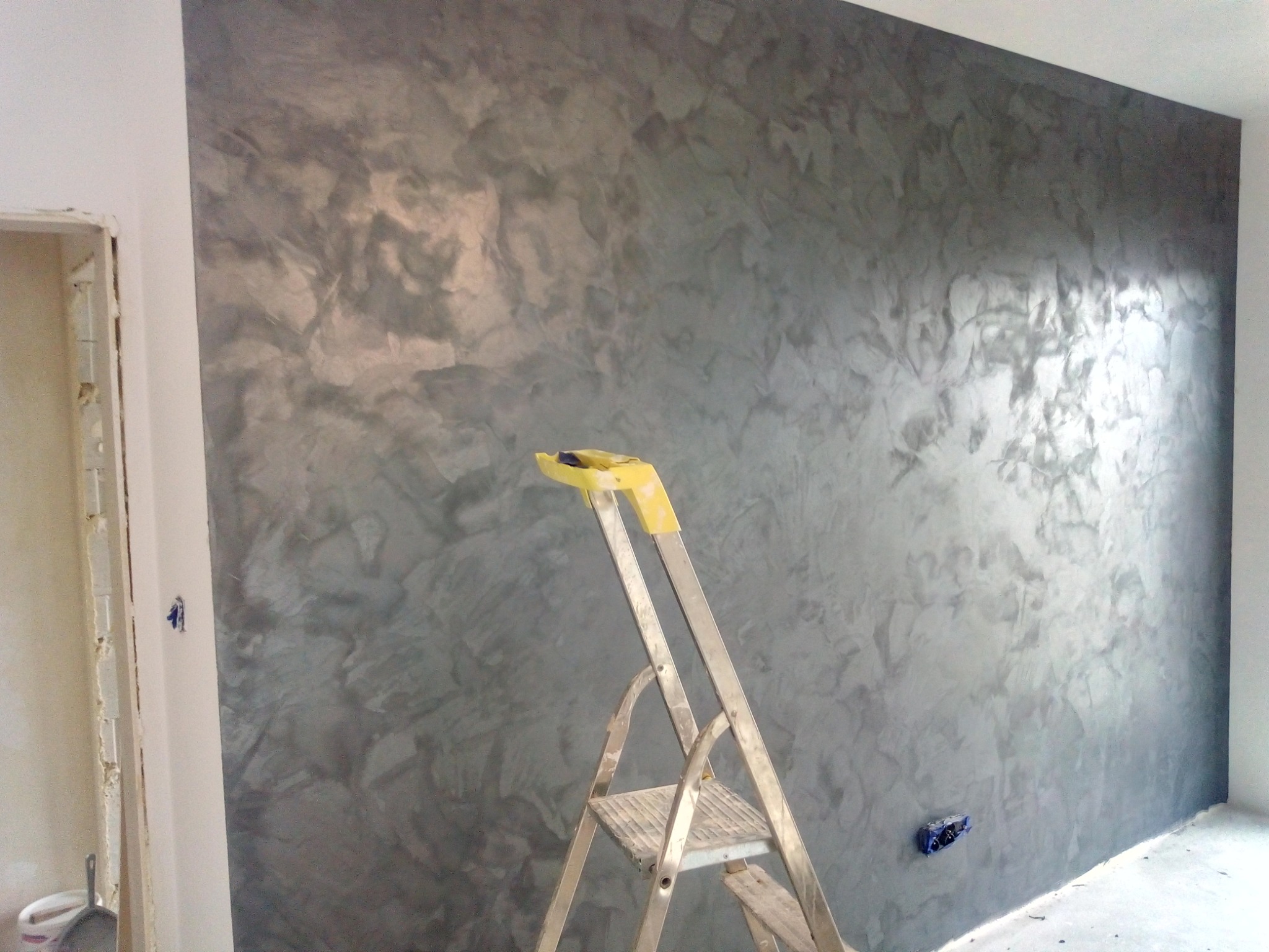 Ściana pokryta dekoracyjną, szarą farbą strukturalną z efektem chmur, widoczna drabina malarska z żółtymi zabezpieczeniami na górze, gniazdko elektryczne zamontowane nisko przy podłodze.