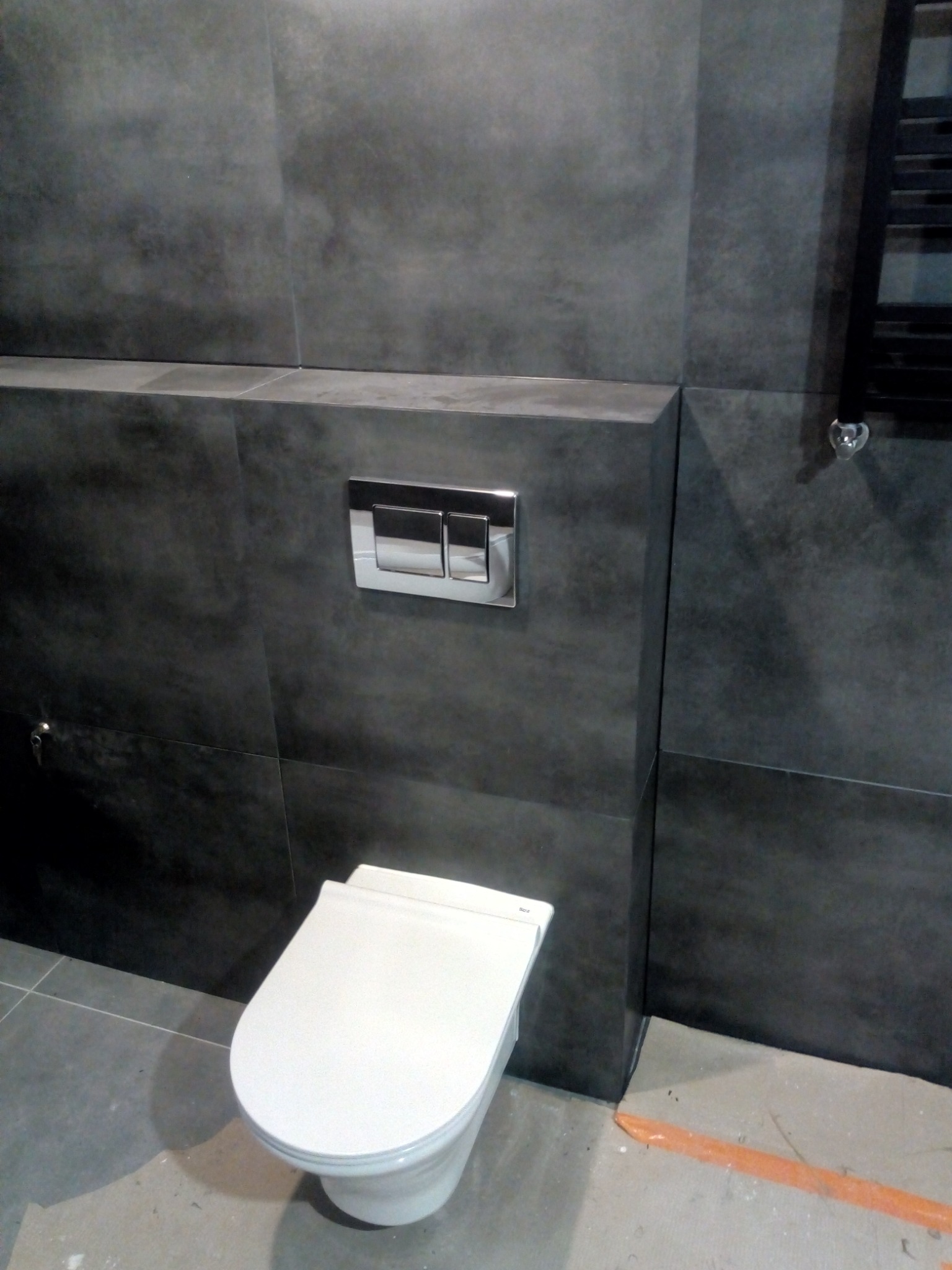 Nowoczesna toaleta podwieszana z chromowanym przyciskiem spłukującym, otoczona szarymi płytkami imitującymi beton, z widocznym fragmentem grzejnika drabinkowego i pomarańczową taśmą malarską...