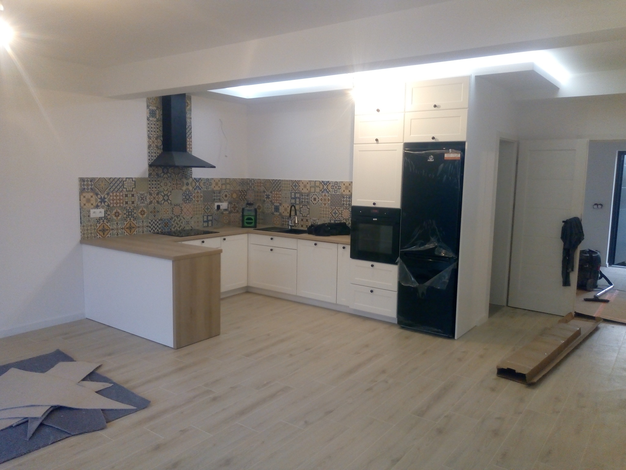 Nowoczesna kuchnia z białymi szafkami, czarną lodówką i wzorzystą mozaiką nad blatem, widoczne kartony na podłodze wskazujące na trwający remont.