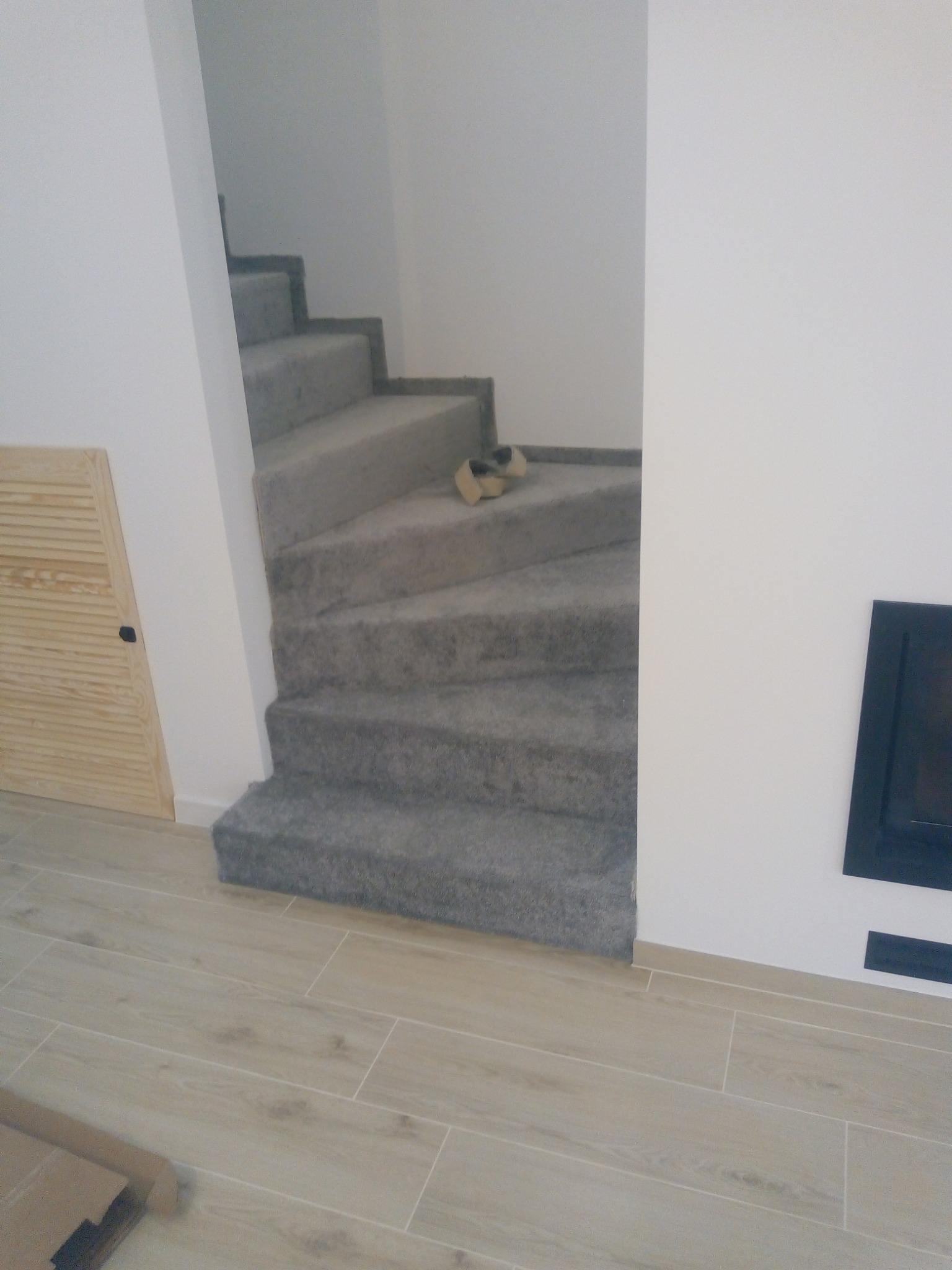 Schody wewnętrzne wyłożone szarym dywanem, z drewnianą szafką w tle i jasnymi płytkami podłogowymi.