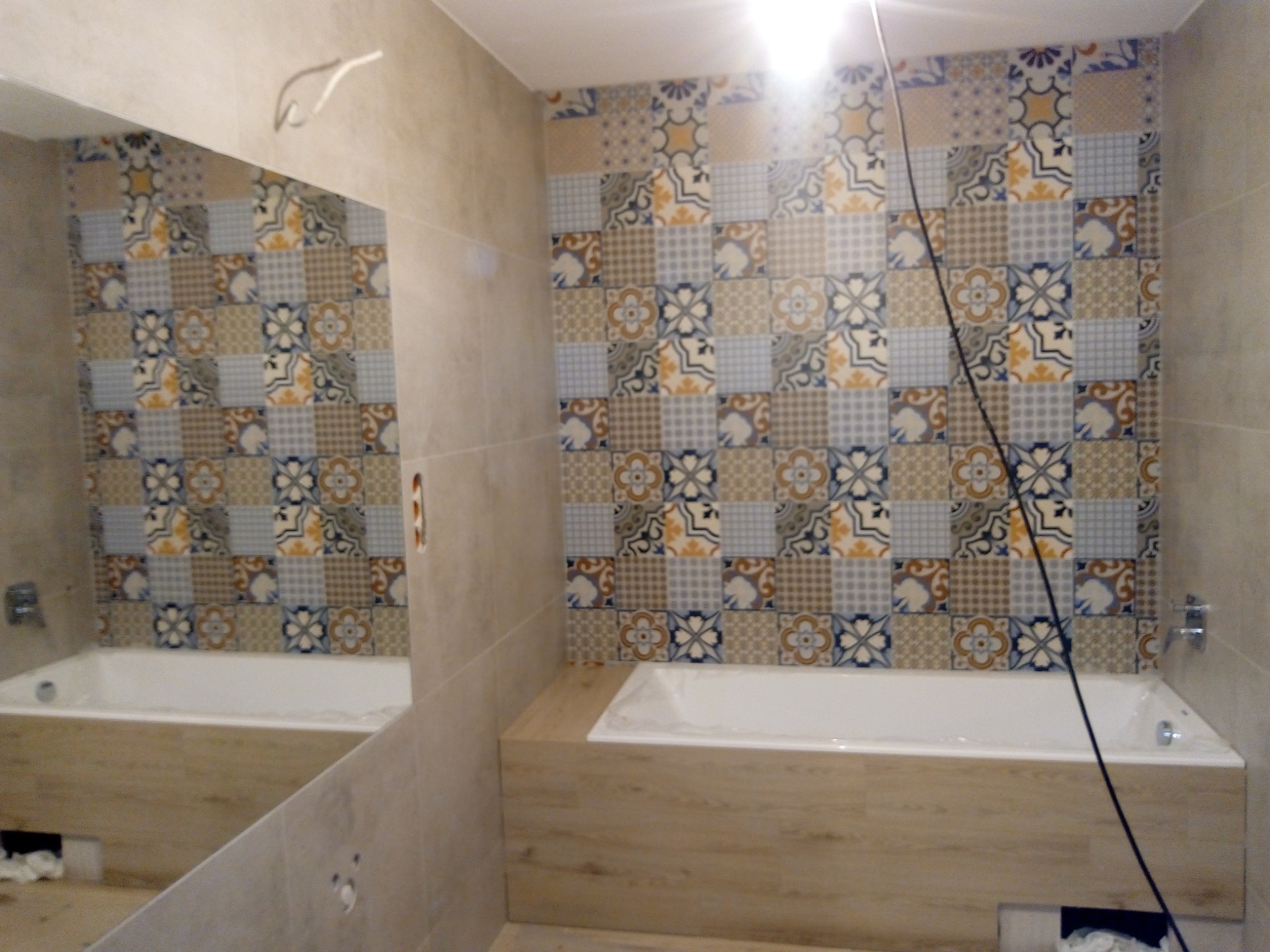 Wykończona łazienka z wanną obudowaną panelami imitującymi drewno i ścianą z płytek w patchworkowy wzór, odbicie w lustrze, widoczne fragmenty instalacji.