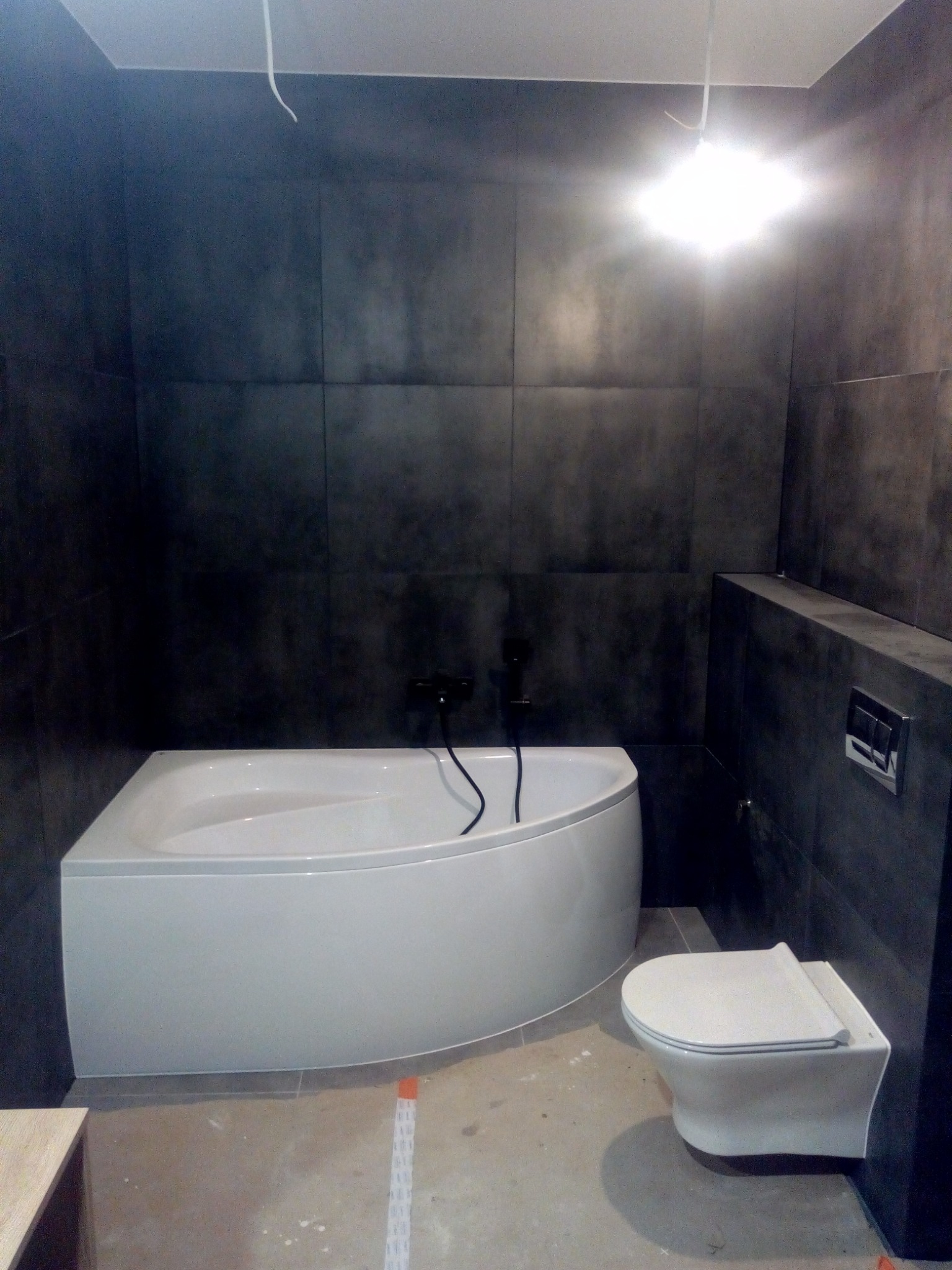 Nowoczesna łazienka w trakcie remontu: biała wanna narożna, wisząca toaleta i duże grafitowe płytki na ścianach, widoczne kable i niedokończona podłoga.