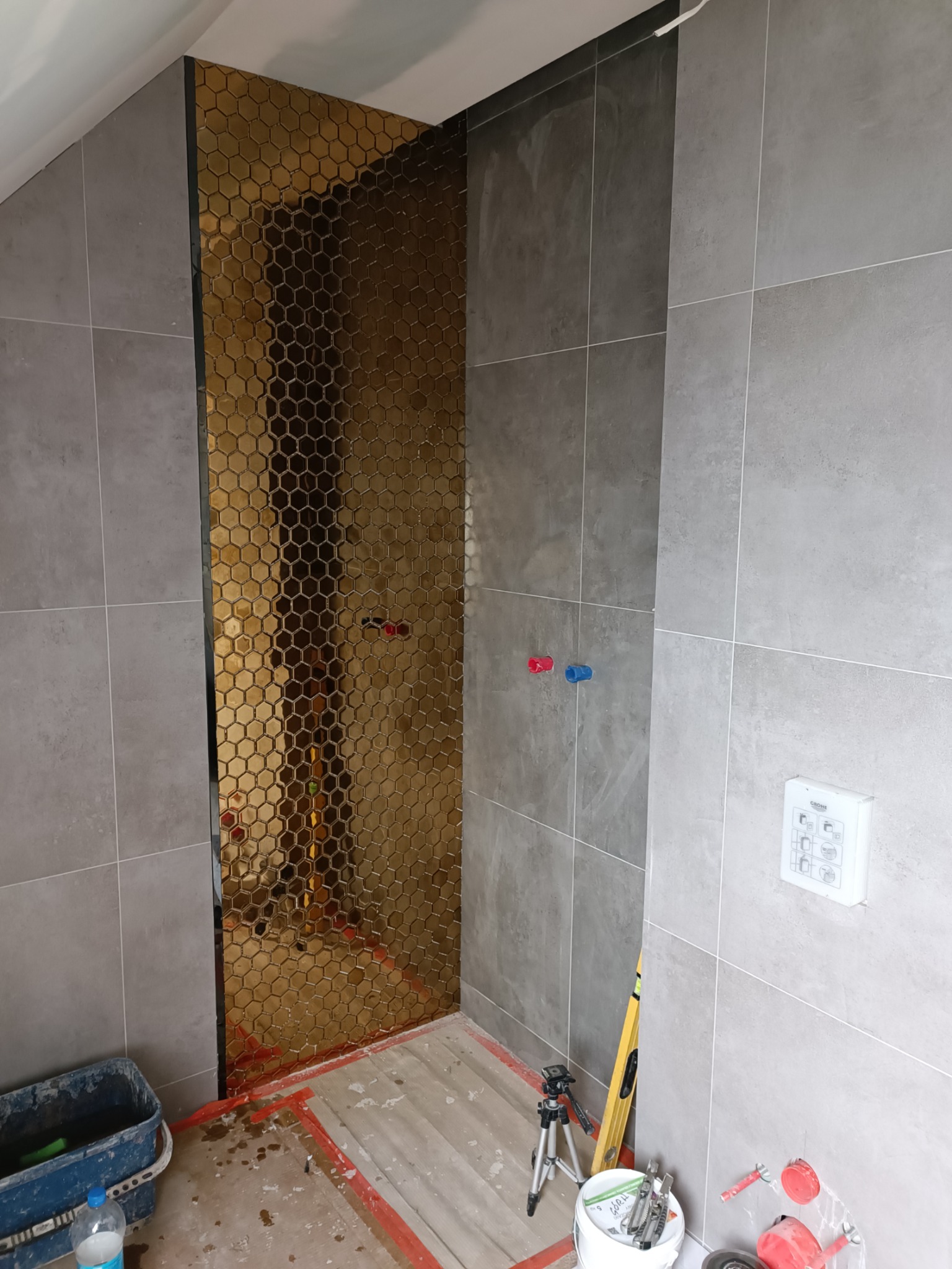 Łazienka w trakcie remontu: ściana wyłożona szarą płytką oraz fragment z mozaiką w kształcie złotych heksagonów, widoczne narzędzia i materiały budowlane.