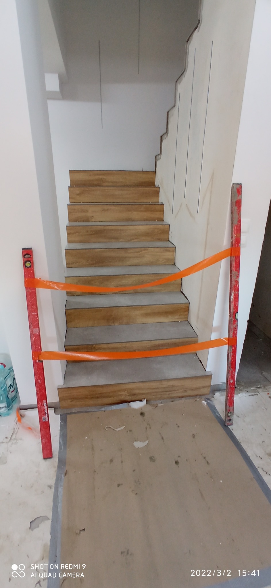 Schody wyłożone płytkami imitującymi drewno, zabezpieczone taśmą ostrzegawczą i poziomnicami, widoczne ślady po pracach wykończeniowych na ścianie.