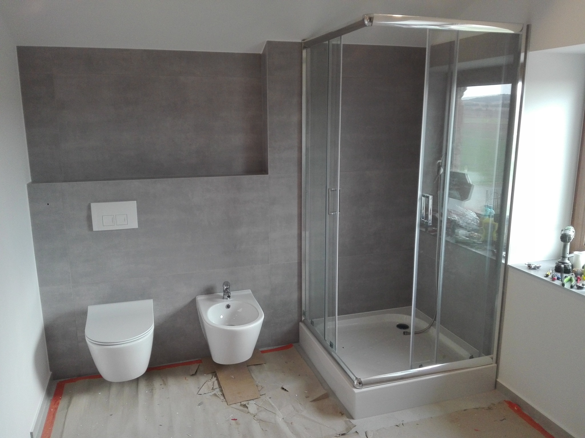 Nowoczesna łazienka w trakcie wykończenia: szare płytki, biała toaleta i bidet, przeszklona kabina prysznicowa, narzędzia na parapecie.