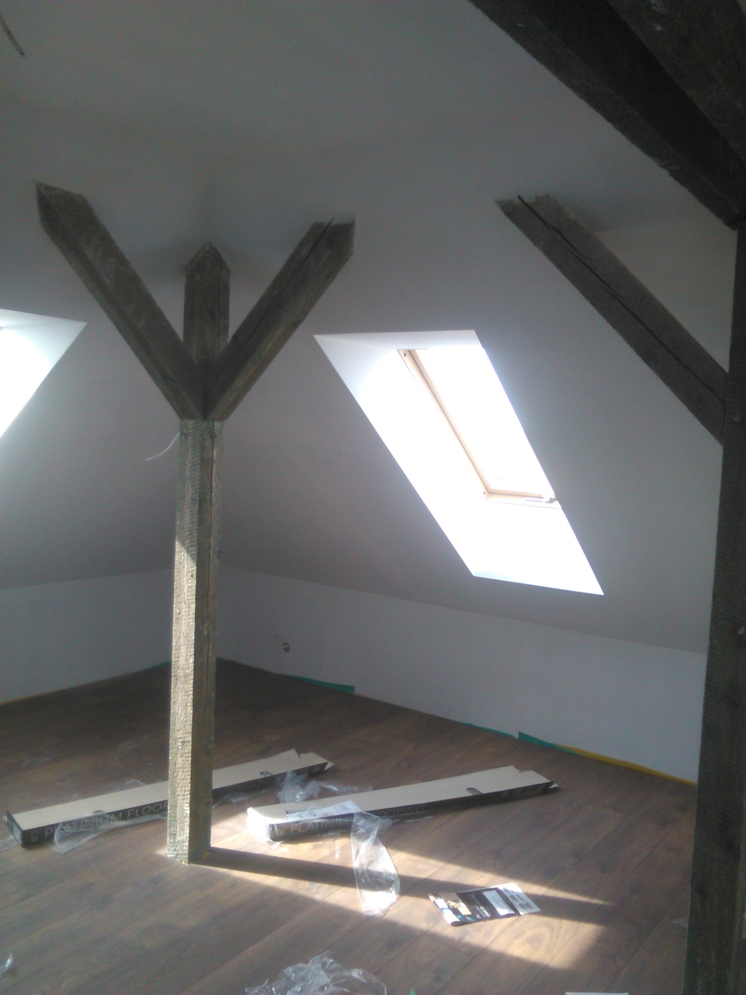 Poddasze z drewnianymi belkami konstrukcyjnymi, oknem dachowym i częściowo ułożoną podłogą z paneli; widoczne opakowania po panelach i ulotka informacyjna producenta.