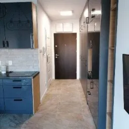 Nowoczesne wykończenie wnętrza mieszkania: szara zabudowa kuchenna z niebieskimi szafkami, korytarz z brązowymi drzwiami i płytkami podłogowymi imitującymi marmur, w tle białe ściany i drzwi.