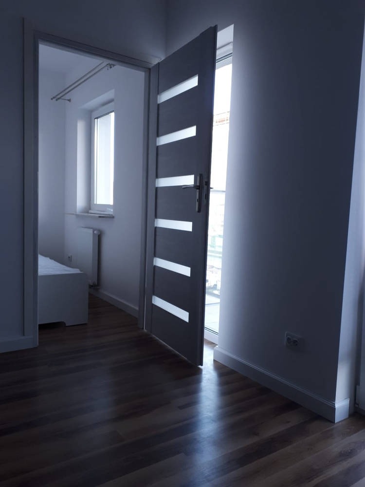 Nowoczesne, grafitowe drzwi wewnętrzne z poziomymi przeszkleniami, uchylone w stronę jasnego pokoju z widocznym łóżkiem i oknem, na tle ciemnej podłogi z paneli.