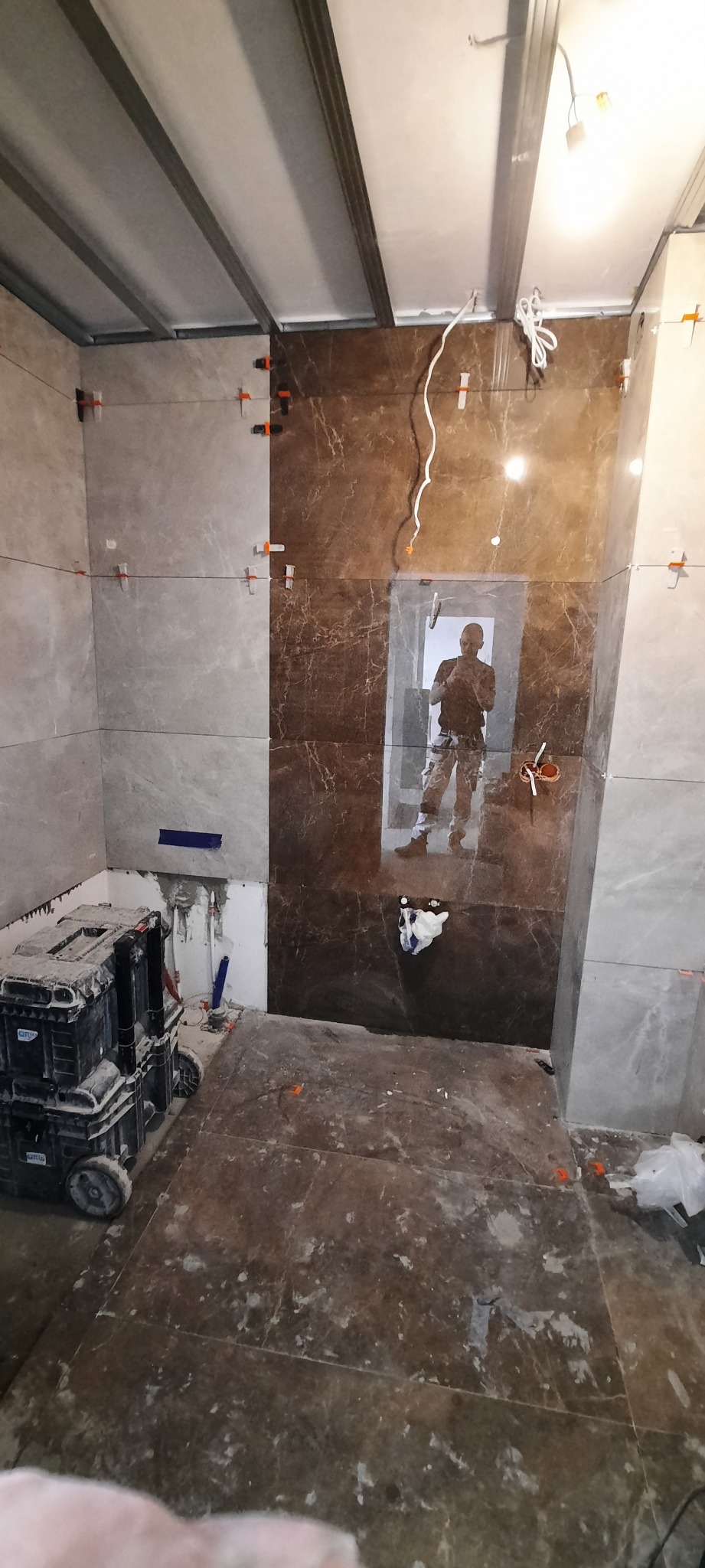 Układanie brązowych i szarych płytek marmuropodobnych w łazience, widoczne krzyżyki dystansowe, odblask sylwetki glazurnika w lustrze z płytek, oraz skrzynki narzędziowe