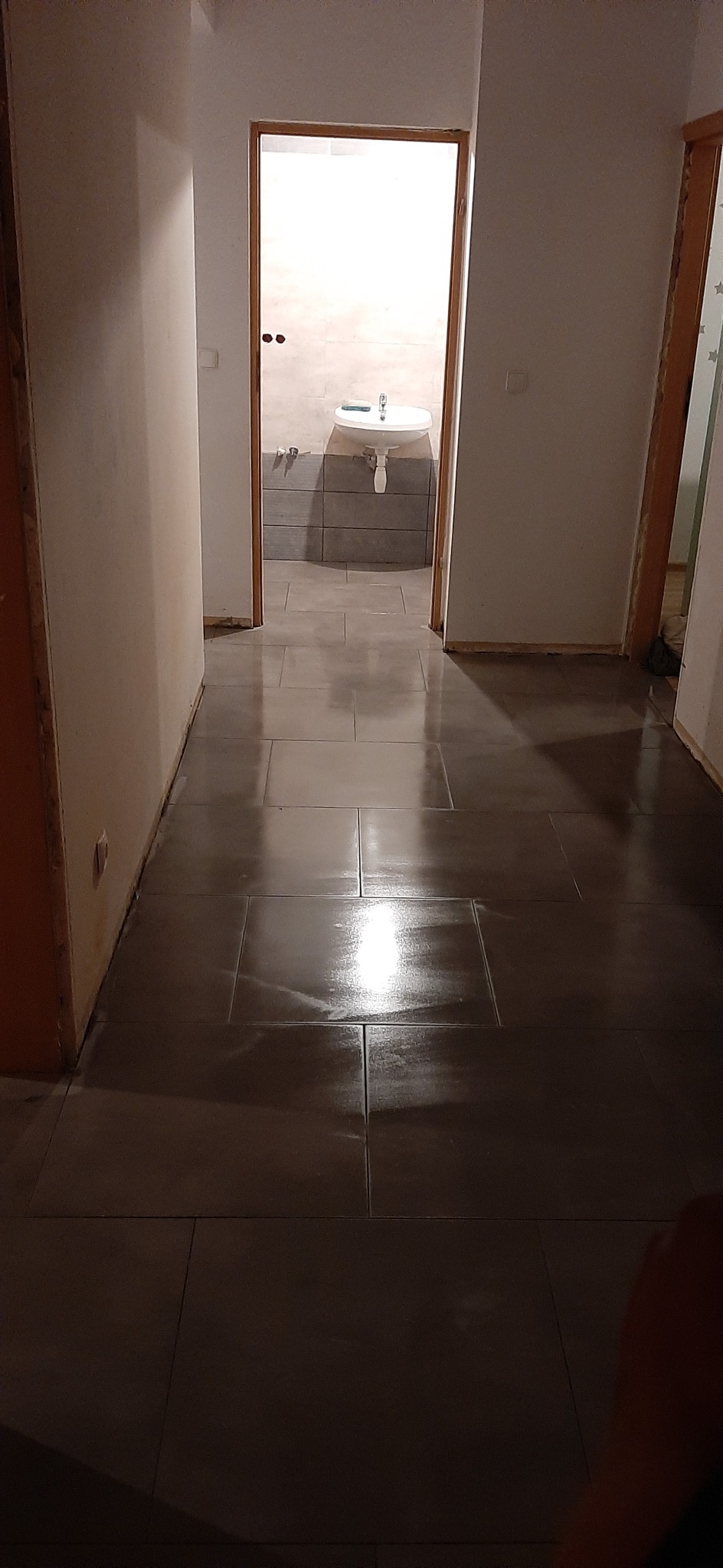 Korytarz z nową, ciemną podłogą z płytek ceramicznych, widok na łazienkę z umywalką na postumencie i szarą glazurą ścienną w tle.