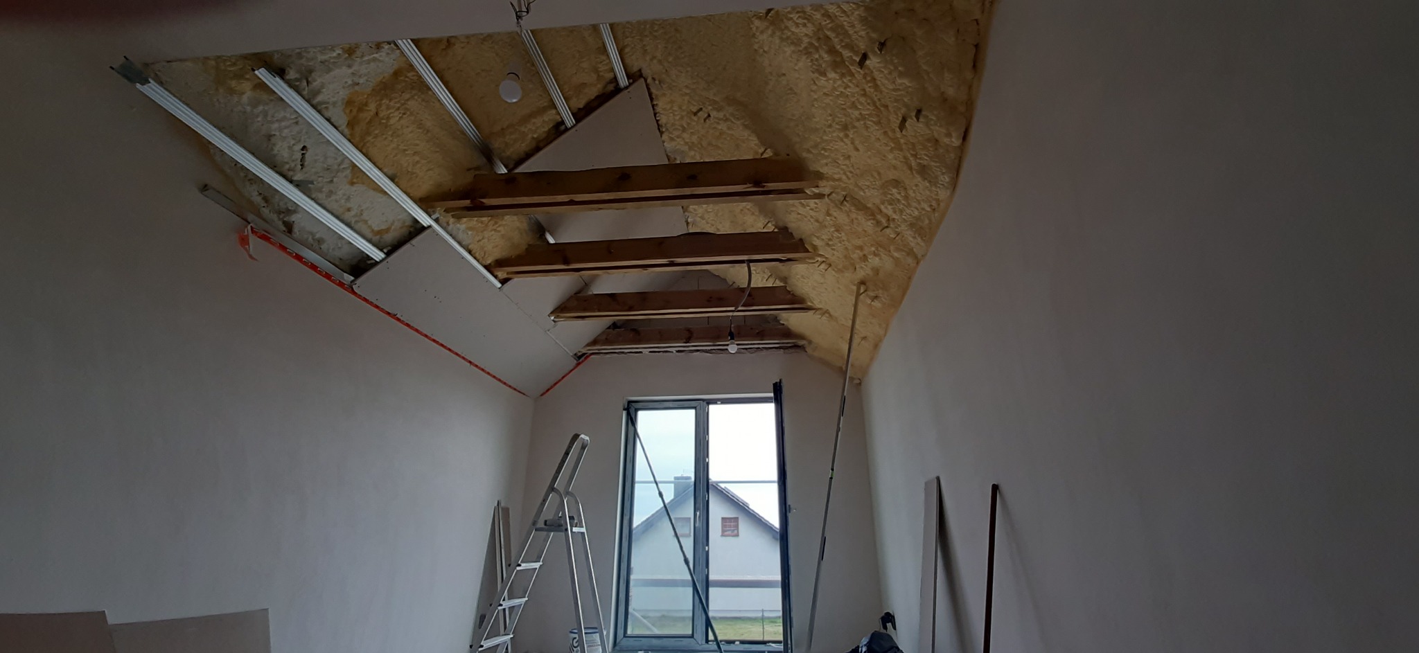 Poddasze w trakcie adaptacji w Poznaniu: widoczne ocieplenie pianą, metalowy stelaż, częściowo zamontowane płyty gipsowo-kartonowe, okno i drabina.