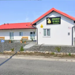 Biały budynek z czerwonym dachem, tablicą informacyjną 'Apartamenty pod Gruszą' i numerem 57, otoczony kamieniami i zielenią.