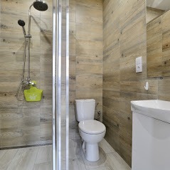 Nowoczesna łazienka z kabiną prysznicową typu walk-in, wykończona płytkami imitującymi drewno, biała ceramika sanitarna, zielony organizer na kosmetyki pod prysznicem.
