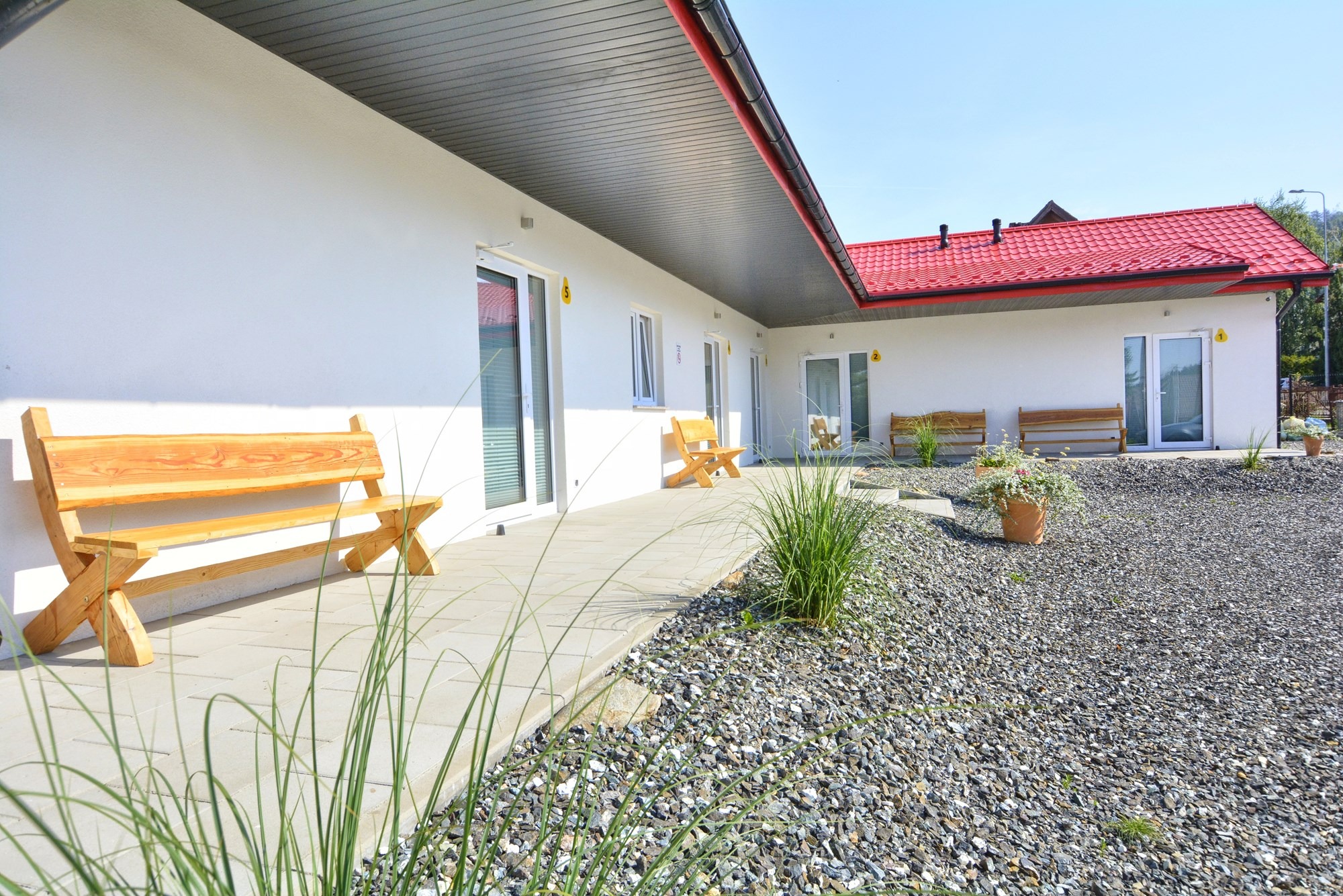 Biały budynek z czerwonym dachem, drewniane ławki przed wejściem, teren wysypany drobnymi kamieniami.