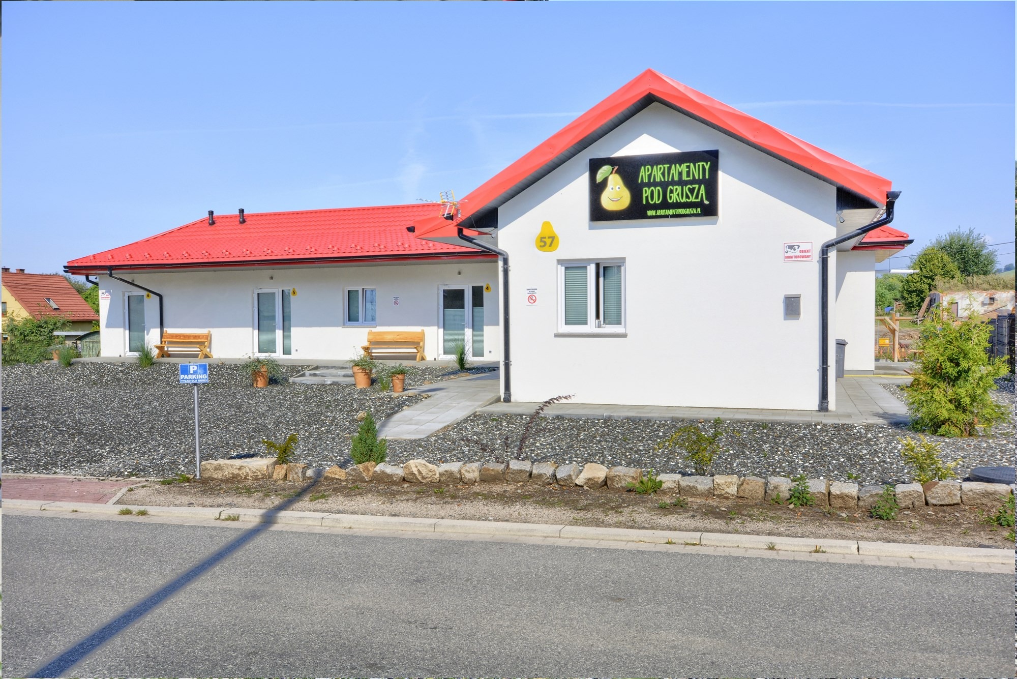 Biały budynek z czerwonym dachem, tablicą informacyjną 'Apartamenty pod Gruszą' i numerem 57, otoczony kamieniami i zielenią.