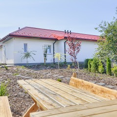 Nowoczesny, parterowy dom z czerwonym dachem widoczny z perspektywy drewnianego stołu piknikowego w ogrodzie, z młodymi drzewkami i korą rozsypaną na ziemi.