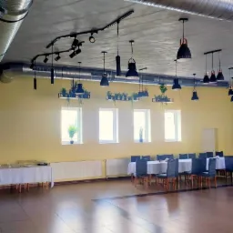 Duża sala z widocznymi przewodami wentylacyjnymi pod sufitem, stoły nakryte białymi obrusami, wiszące lampy i rośliny doniczkowe w oknach.