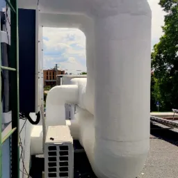 Zewnętrzna jednostka klimatyzacji na dachu, z widocznymi grubymi, zaizolowanymi rurami, na tle błękitnego nieba z chmurami.