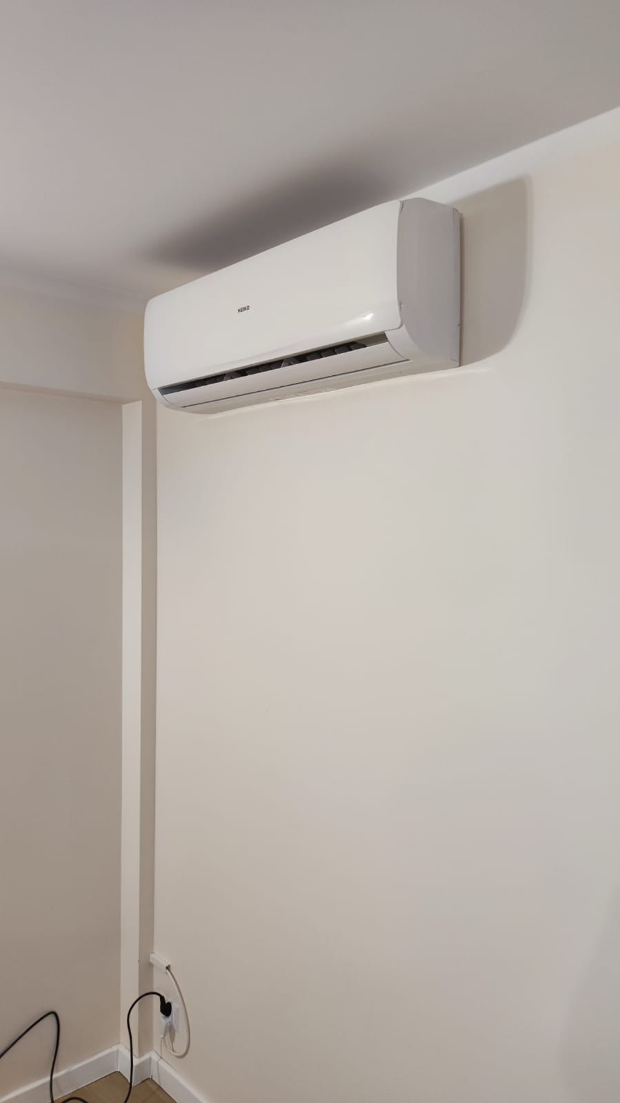 Biały klimatyzator Heko zamontowany na jasnej ścianie w pomieszczeniu, widoczna instalacja elektryczna z wtyczką w gniazdku.
