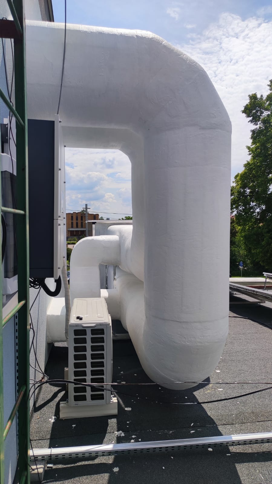 Białe, zaizolowane rury wentylacyjne na dachu budynku, obok jednostka zewnętrzna klimatyzacji, z widokiem na budynek w tle.