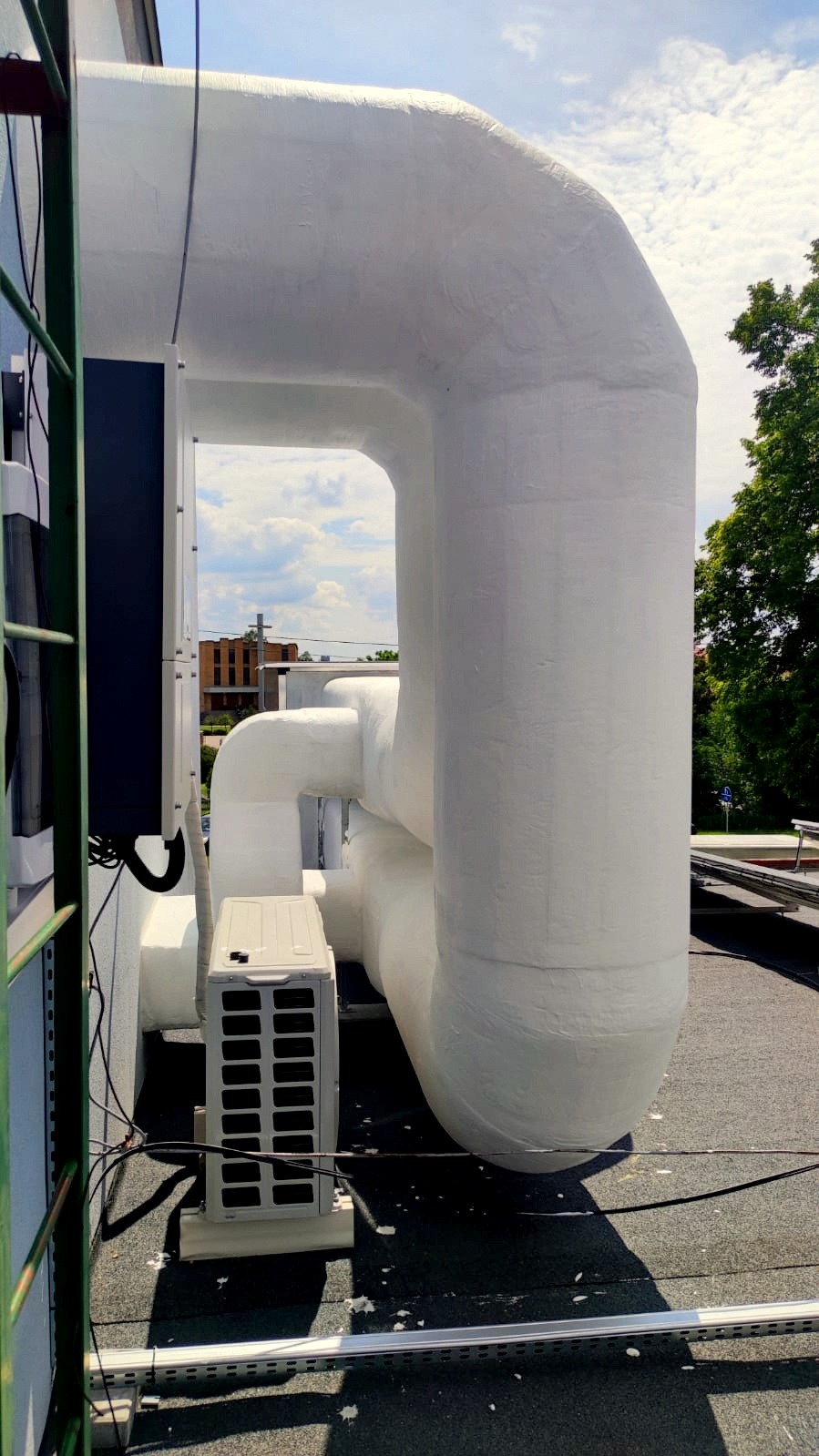 Zewnętrzna jednostka klimatyzacji na dachu, z widocznymi grubymi, zaizolowanymi rurami, na tle błękitnego nieba z chmurami.