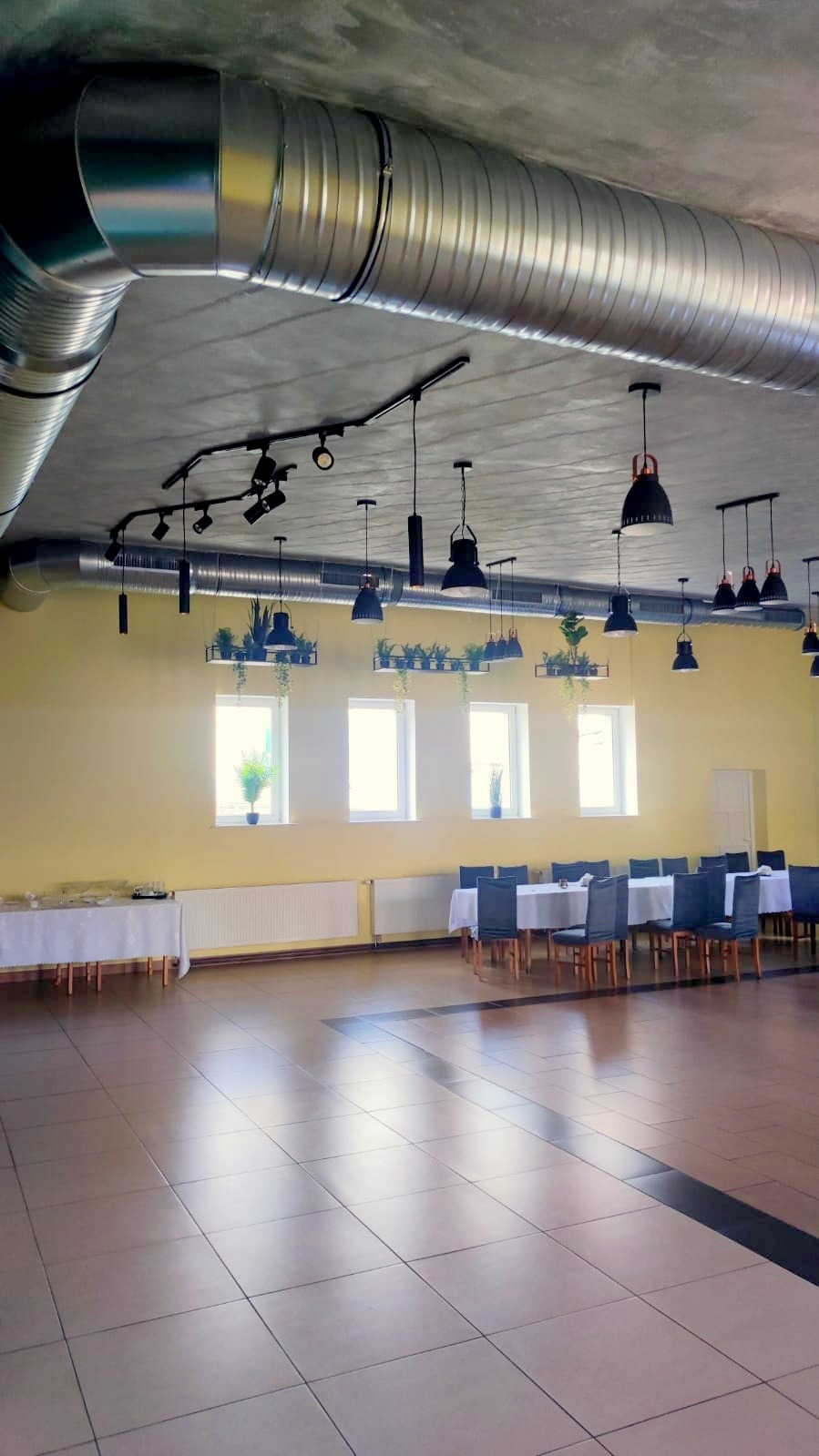 Duża sala z widocznym systemem wentylacyjnym pod sufitem, z rurami rozprowadzonymi po całej przestrzeni, oświetleniem punktowym i wiszącymi lampami, stołami przygotowanymi do przyjęcia gości.
