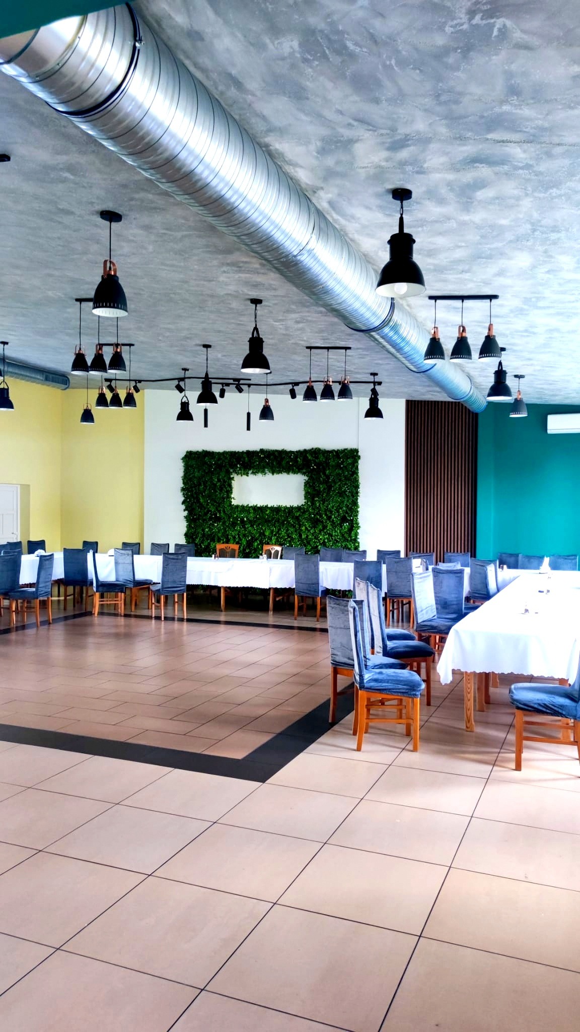 Wnętrze sali bankietowej z widocznym systemem wentylacyjnym pod sufitem, oświetleniem punktowym i aranżacją stołów z krzesłami w niebieskich pokrowcach.