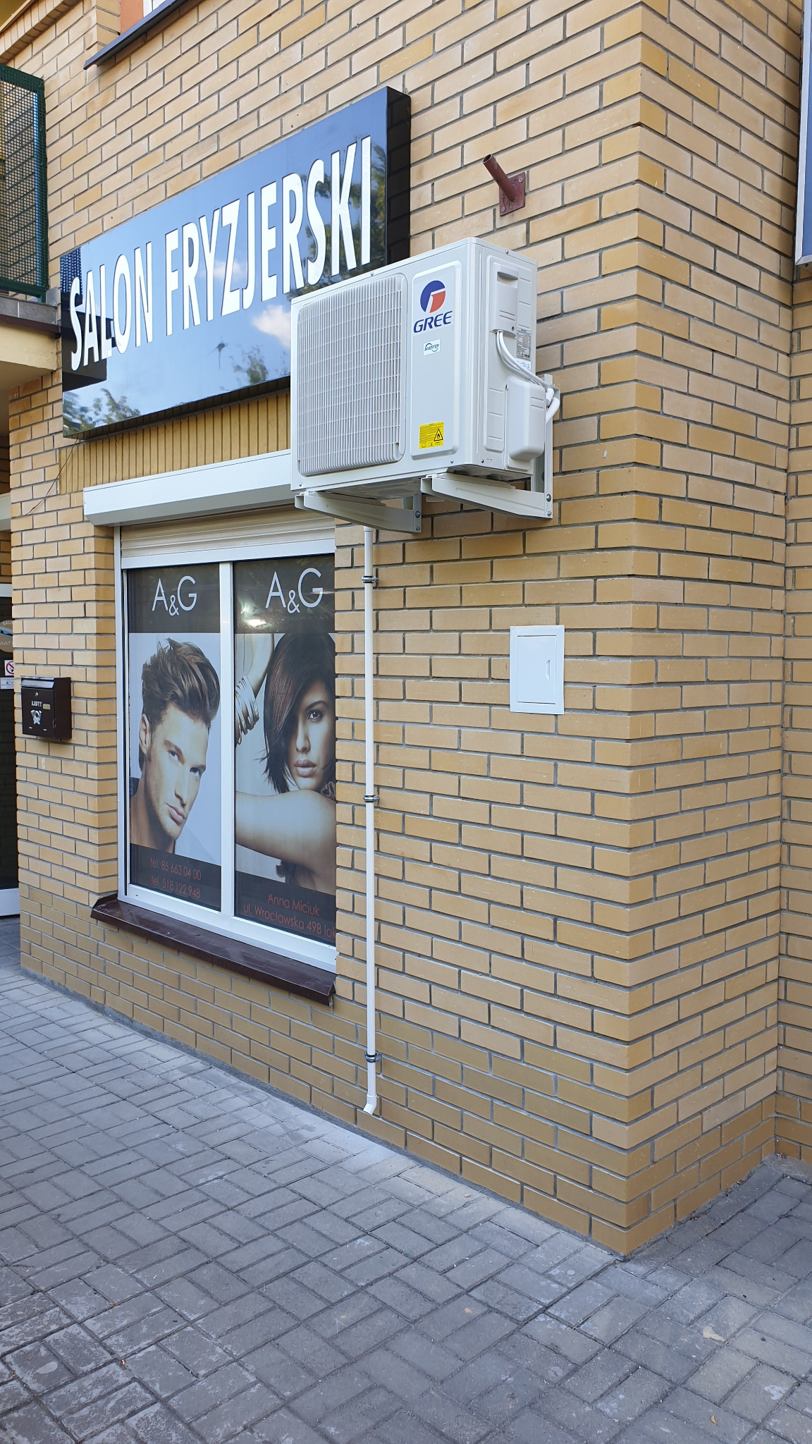 Zewnętrzna jednostka klimatyzacji Gree zamontowana na ścianie budynku salonu fryzjerskiego z cegły klinkierowej, widoczne rury instalacyjne i szyld salonu.