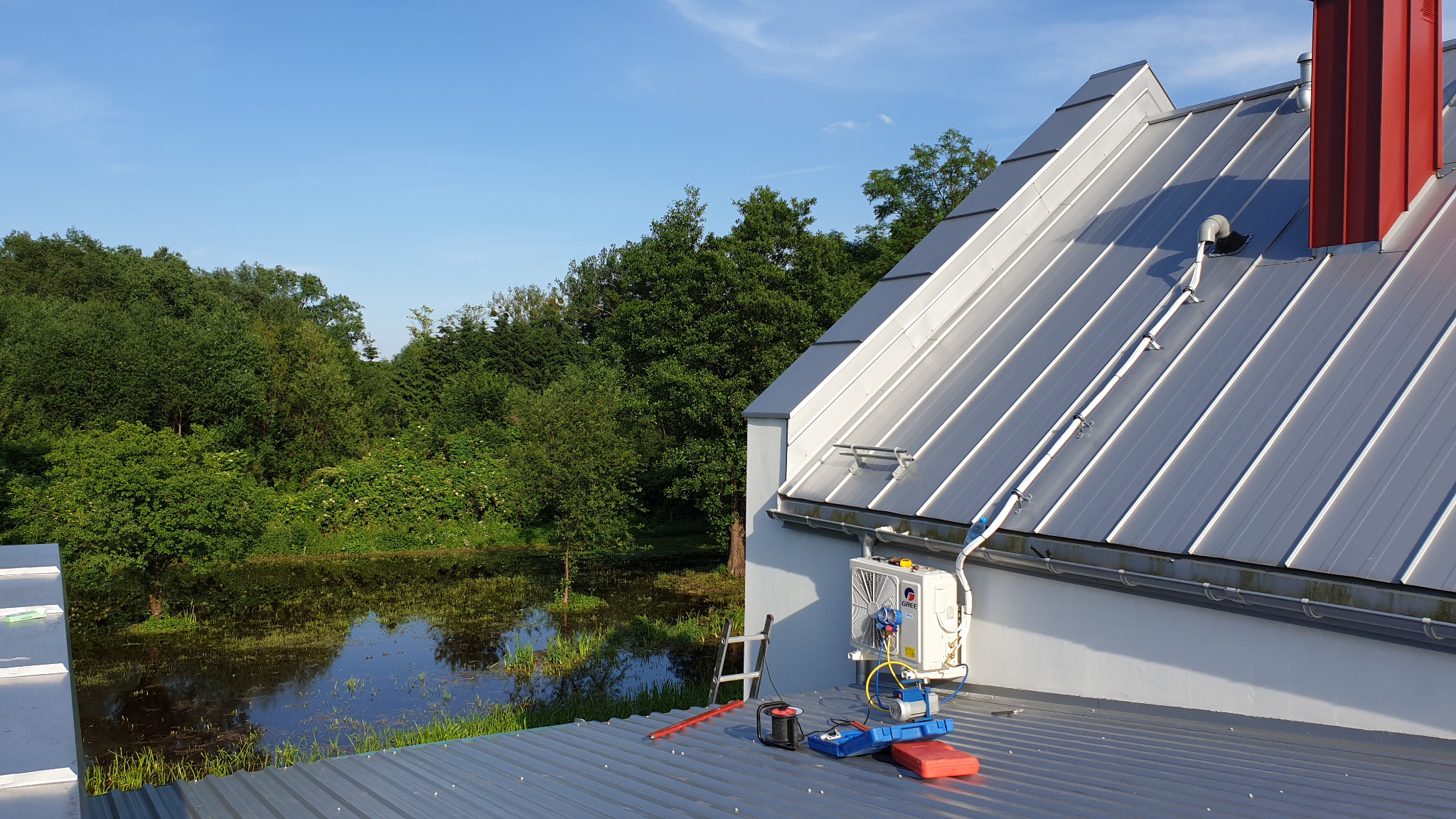 Montaż jednostki zewnętrznej klimatyzacji na dachu domu z widokiem na zarośnięty staw. Widoczne narzędzia pracy, drabina oparta o ścianę i przewody odprowadzające skropliny.