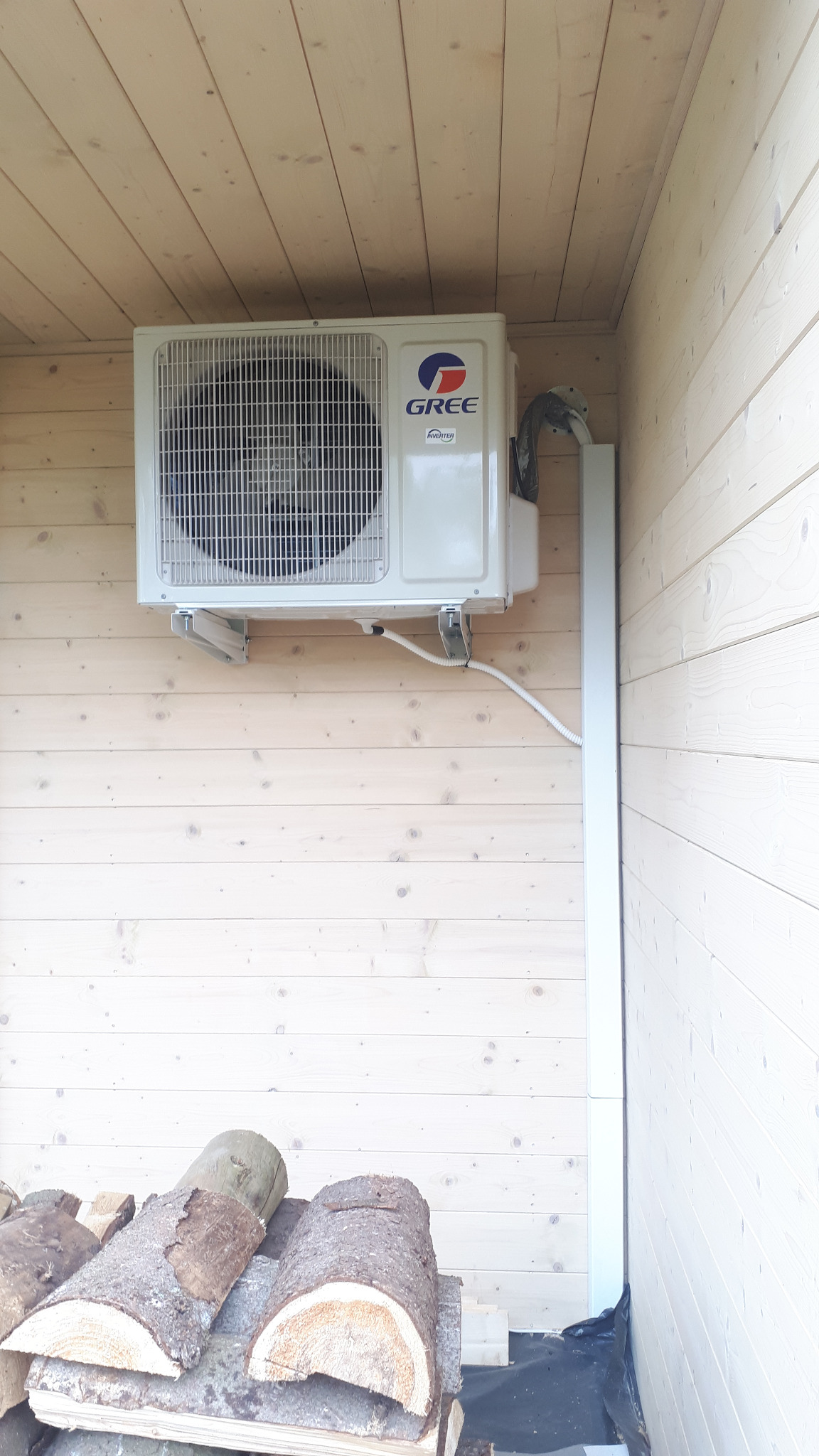 Zewnętrzna jednostka klimatyzacji marki Gree zamontowana na drewnianej ścianie domku, widoczne przewody w białej listwie i stos drewna opałowego u dołu.