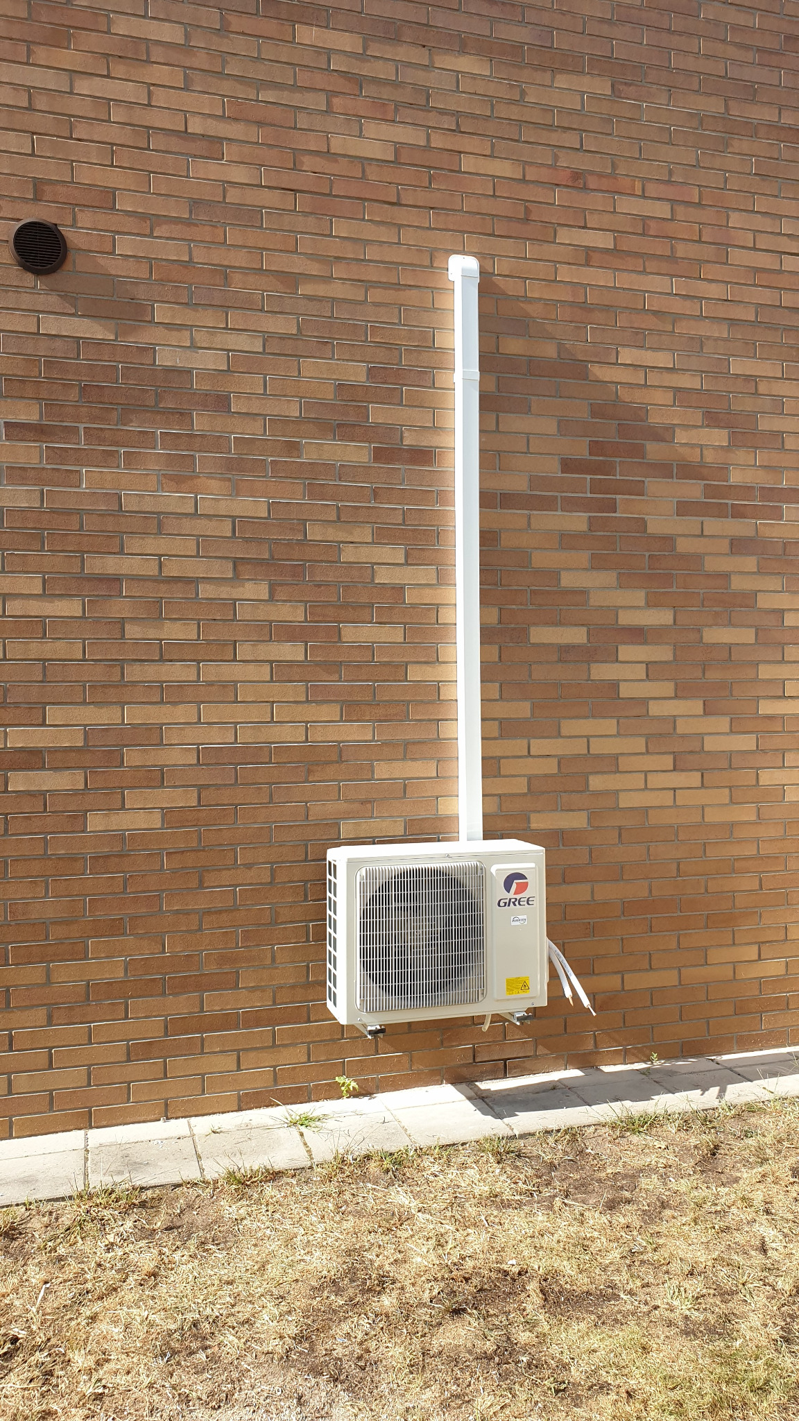 Zamontowana jednostka zewnętrzna klimatyzacji Gree na ceglanej ścianie budynku, z widoczną rurą odprowadzającą skropliny i fragmentem trawnika u dołu kadru.
