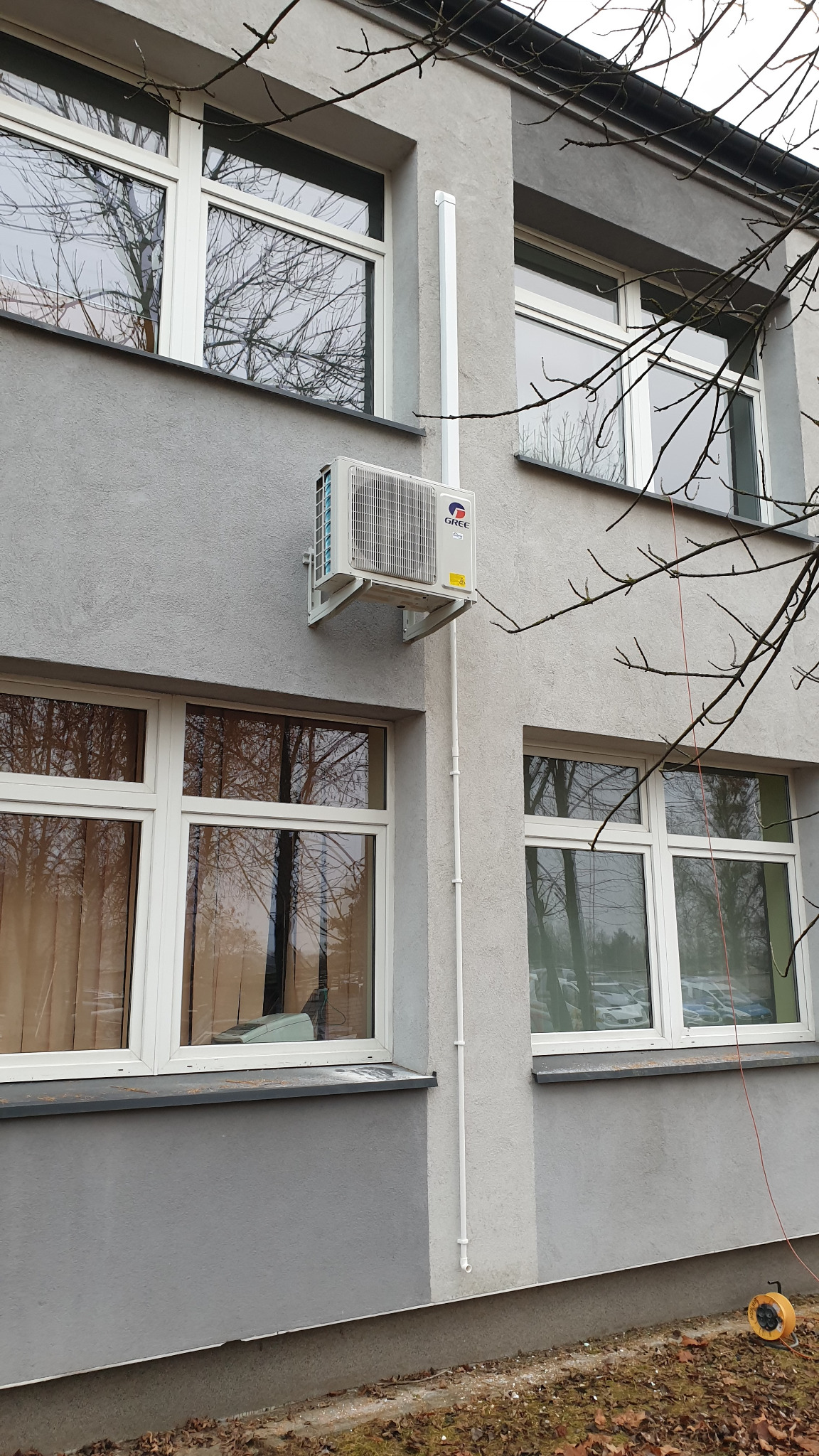Zewnętrzna jednostka klimatyzacji marki Gree zamontowana na szarej ścianie budynku z widocznymi oknami i instalacją odprowadzającą skropliny. Na dole widoczna zwinięta przedłużka elektryczna.