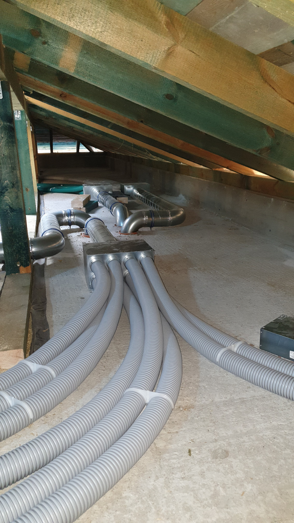 Instalacja przewodów wentylacyjnych w przestrzeni poddasza w Białymstoku, widoczne szare, karbowane rury oraz metalowe elementy systemu wentylacyjnego.