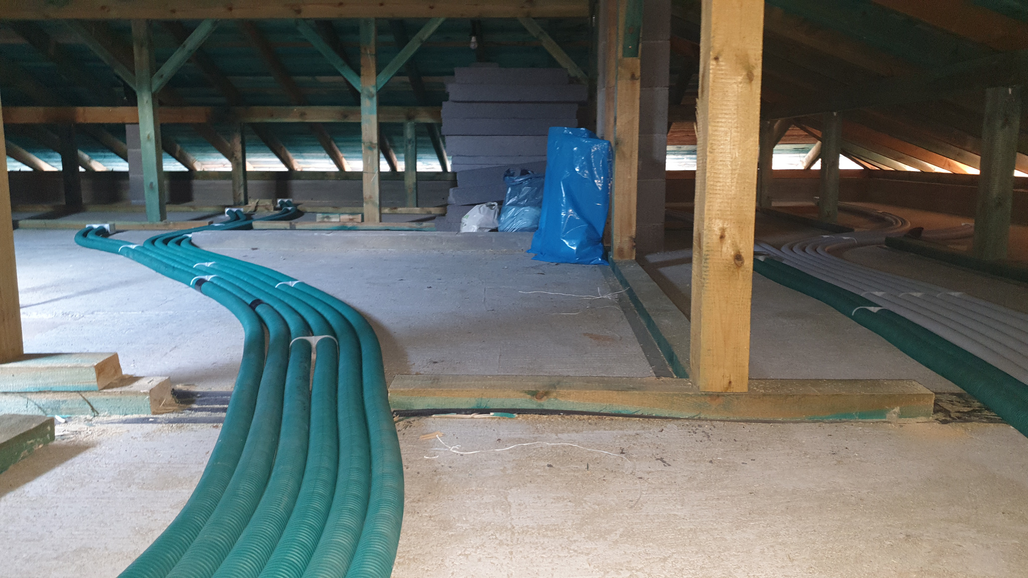 Instalacja przewodów wentylacyjnych na poddaszu, widoczne rury w kolorze zielonym i szarym, drewniana konstrukcja dachu i izolacja termiczna w tle.