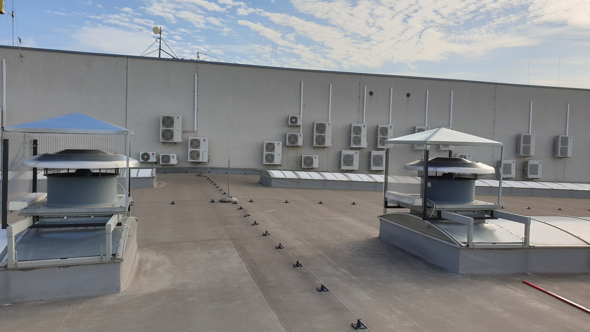 Widok dachu budynku z zainstalowanymi jednostkami zewnętrznymi klimatyzatorów i systemami wentylacyjnymi z metalowymi daszkami ochronnymi.