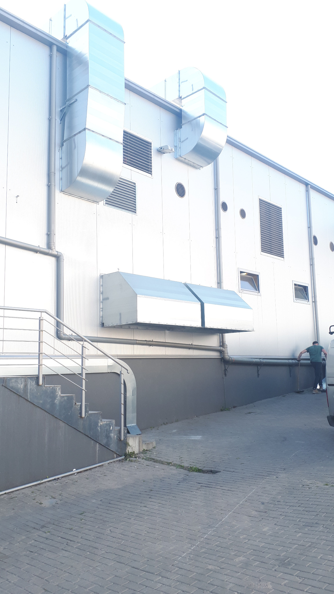 Zewnętrzny widok budynku przemysłowego z systemem wentylacyjnym i klimatyzacją, widoczne metalowe kanały wentylacyjne, kratki wlotowe i fragment osoby przy samochodzie.