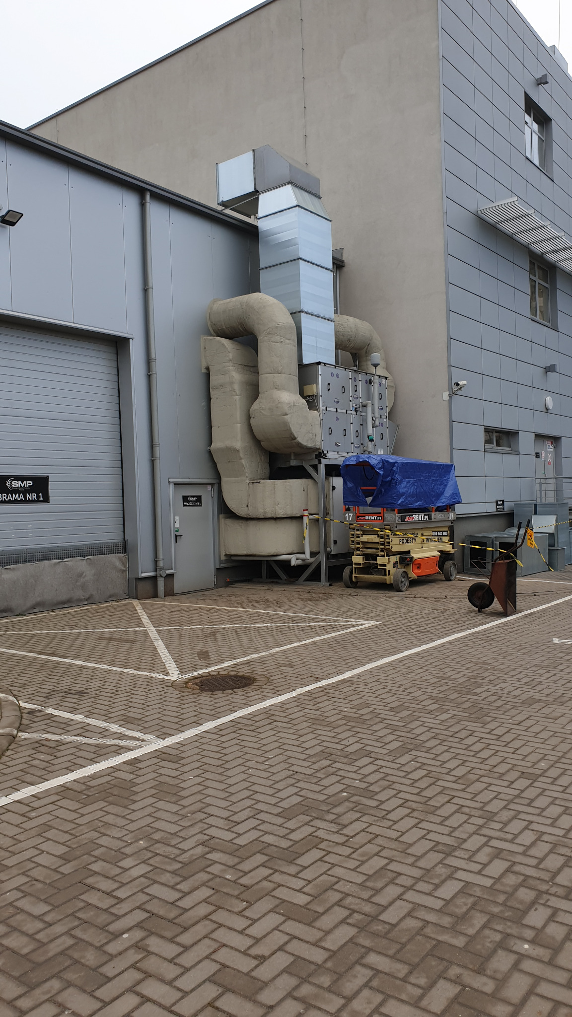 Zewnętrzna instalacja przemysłowa klimatyzacji z rurami i wentylacją przy budynku, platforma robocza pod niebieską plandeką, barierki ostrzegawcze i wózek budowlany.