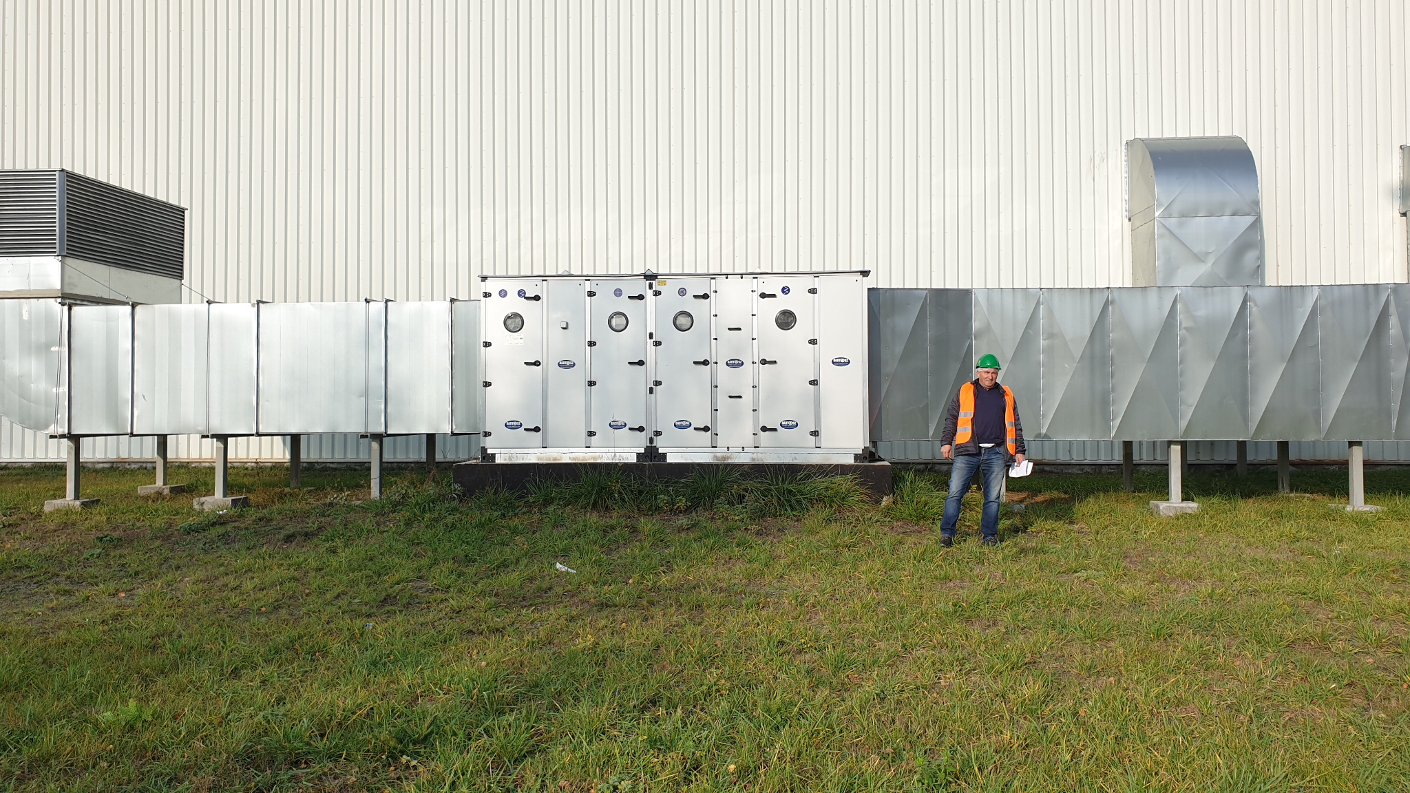 Instalacja przemysłowej centrali wentylacyjnej z kanałami wentylacyjnymi; pracownik w kasku i kamizelce odblaskowej stoi przed instalacją na tle hali.