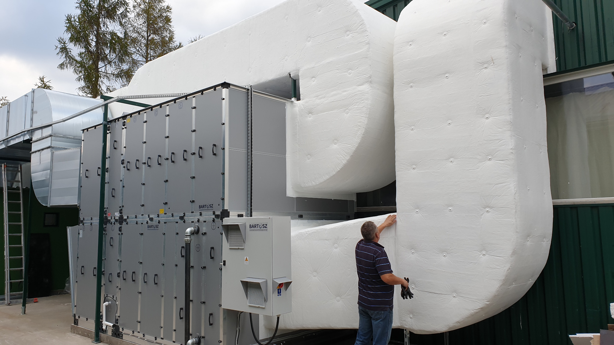 Instalacja przemysłowej wentylacji z izolacją, widoczny mężczyzna w trakcie prac wykończeniowych przy izolacji kanałów wentylacyjnych.