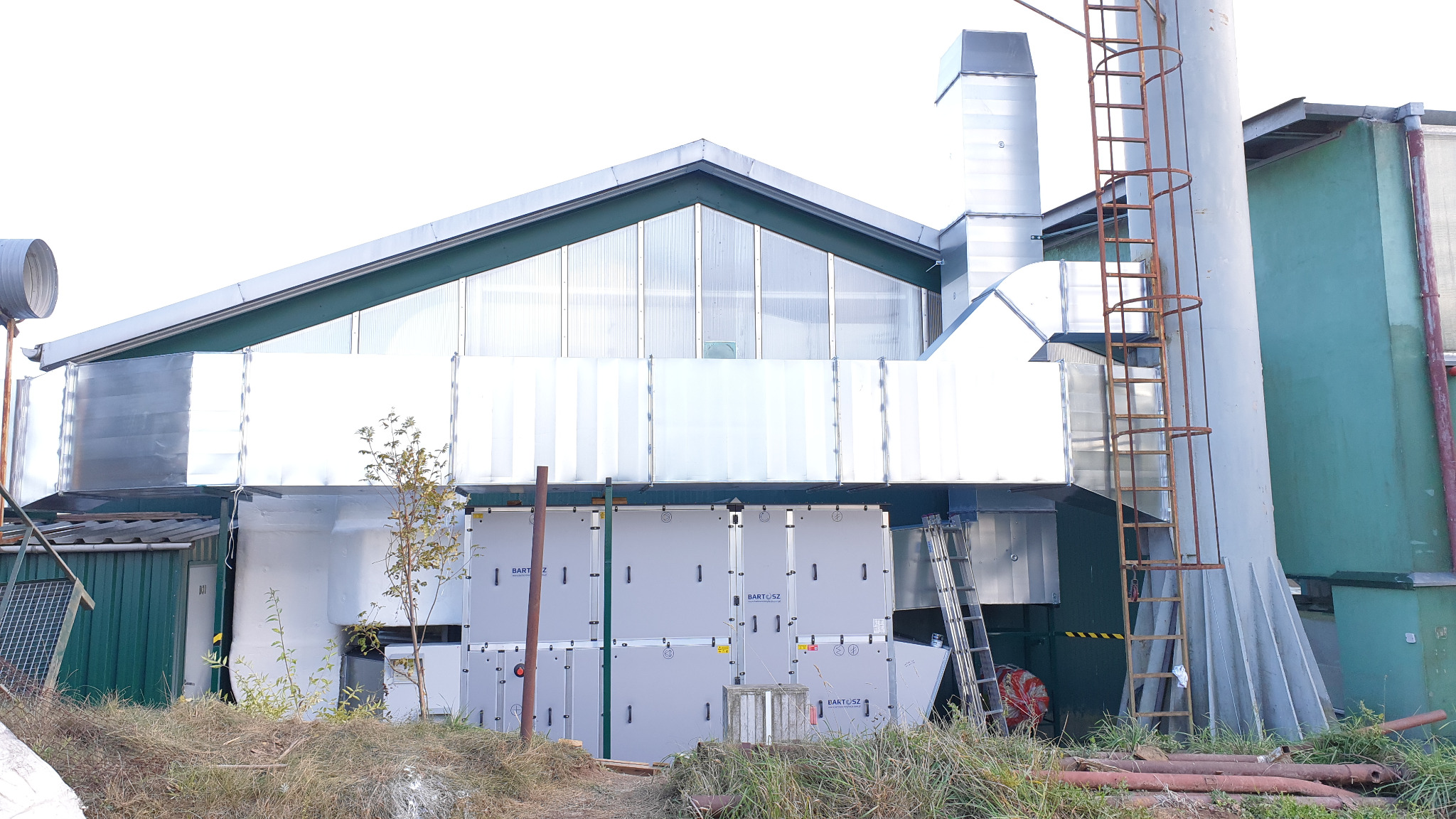 Zewnętrzna instalacja klimatyzacyjna przemysłowa z dużą ilością modułów, widoczne rury wentylacyjne, drabina i fragment budynku przemysłowego.