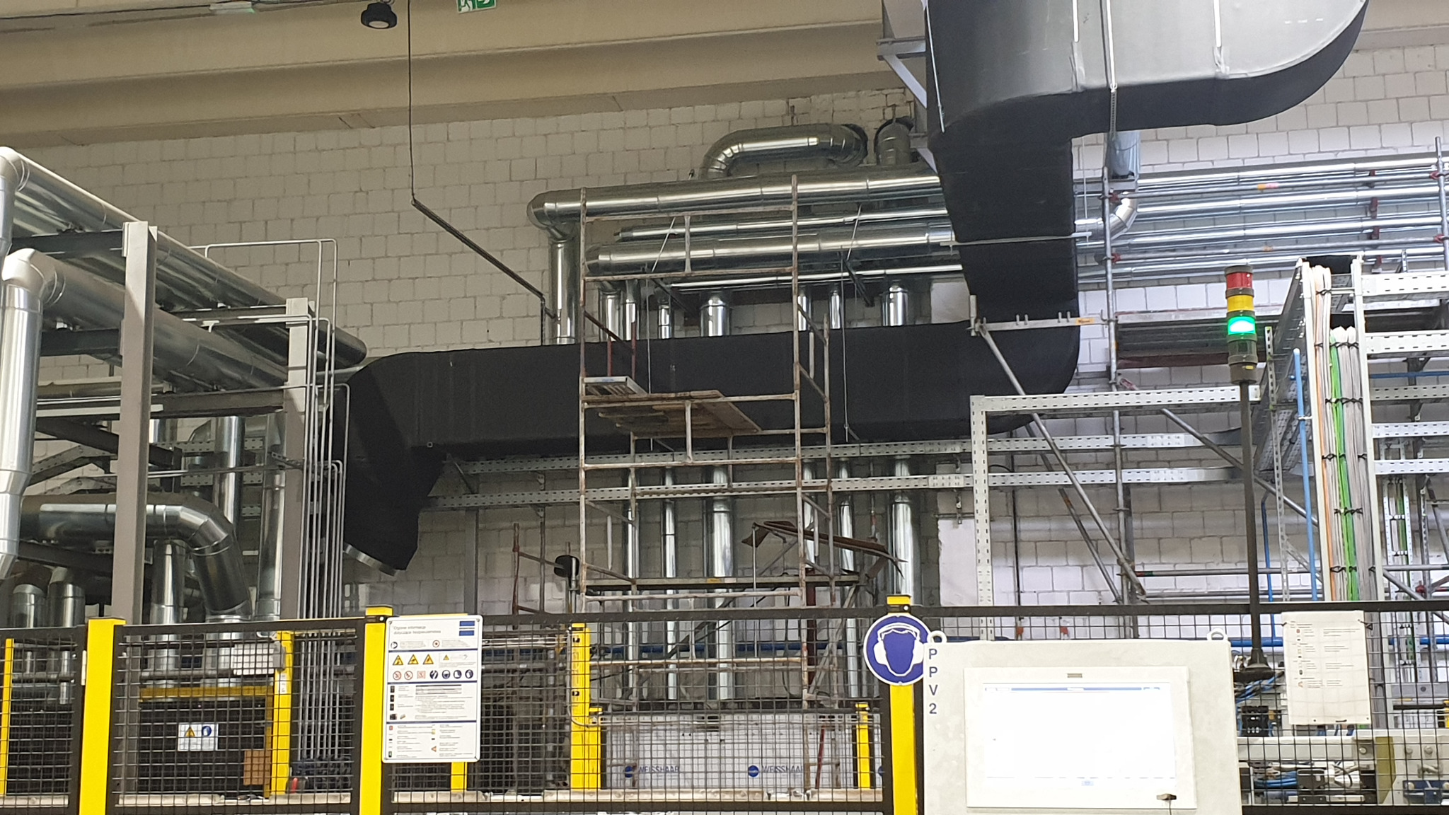 Instalacja przemysłowa z rurami wentylacyjnymi i rusztowaniem na tle ściany z białej cegły, widoczny sygnalizator świetlny z zielonym światłem.