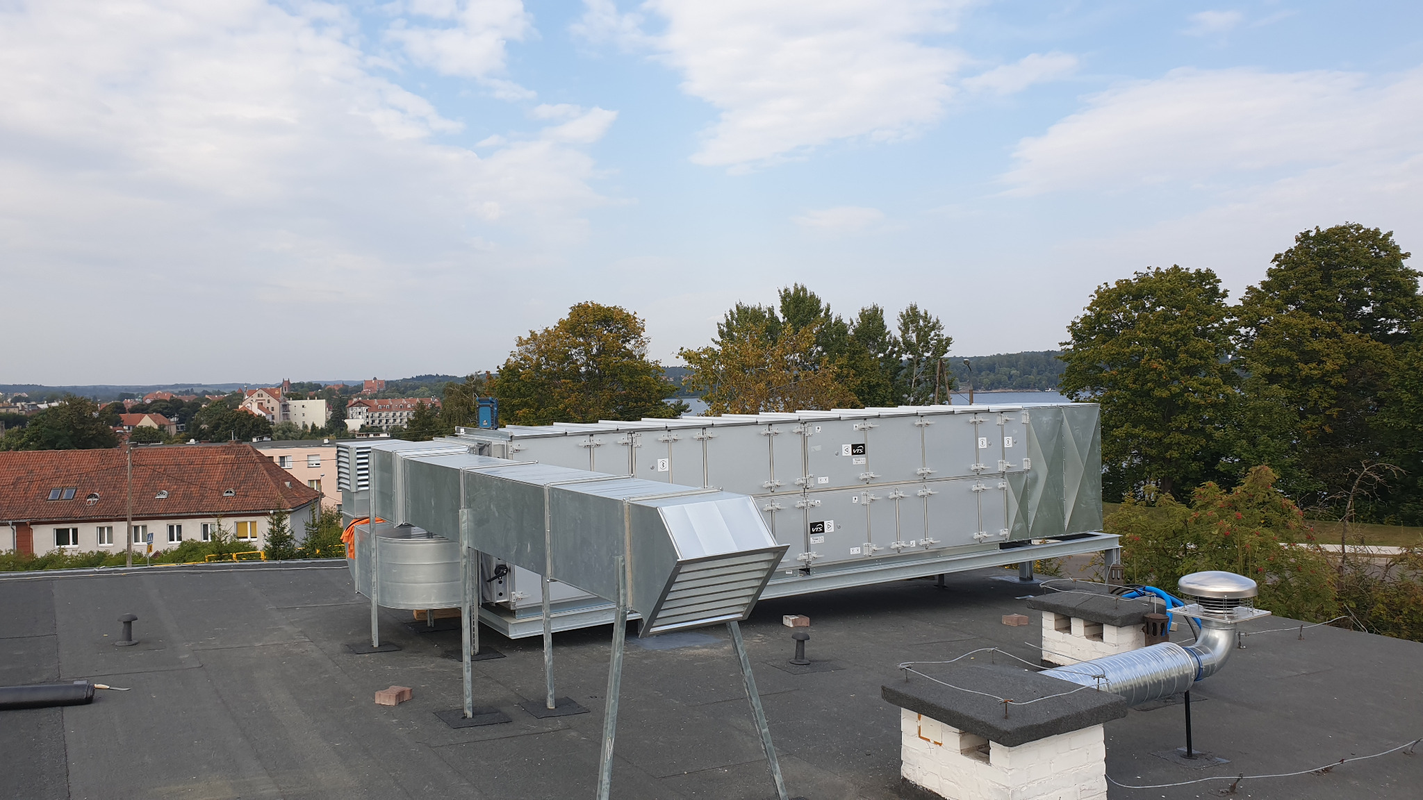 Instalacja wentylacyjna z kanałami na dachu budynku, widok na miasto i jezioro w tle.