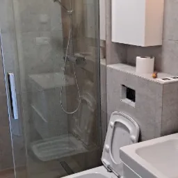 Wykończona łazienka z kabiną prysznicową typu walk-in, podwieszaną toaletą z otwartą klapą, szafką nad toaletą i umywalką z szafką, wszystko w szaro-białej kolorystyce.