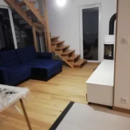 Jasne drewniane schody prowadzące na górę, niebieska sofa, biała szafka RTV z kominkiem w nowoczesnym salonie z drewnianą podłogą.