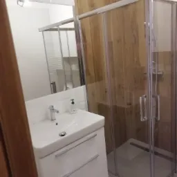 Nowoczesna łazienka z kabiną prysznicową typu walk-in z deszczownicą i umywalką nablatową z szafką w kolorze białym, wykończona płytkami imitującymi drewno.