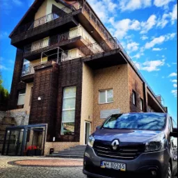 Szary bus Renault Trafic parkuje przed hotelem o drewnianej elewacji i balkonami, błękitne niebo z chmurami w tle.