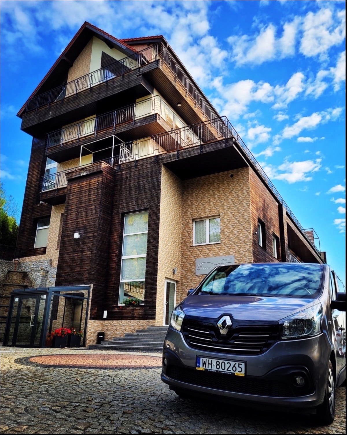 Szary bus Renault Trafic parkuje przed hotelem o drewnianej elewacji i balkonami, błękitne niebo z chmurami w tle.
